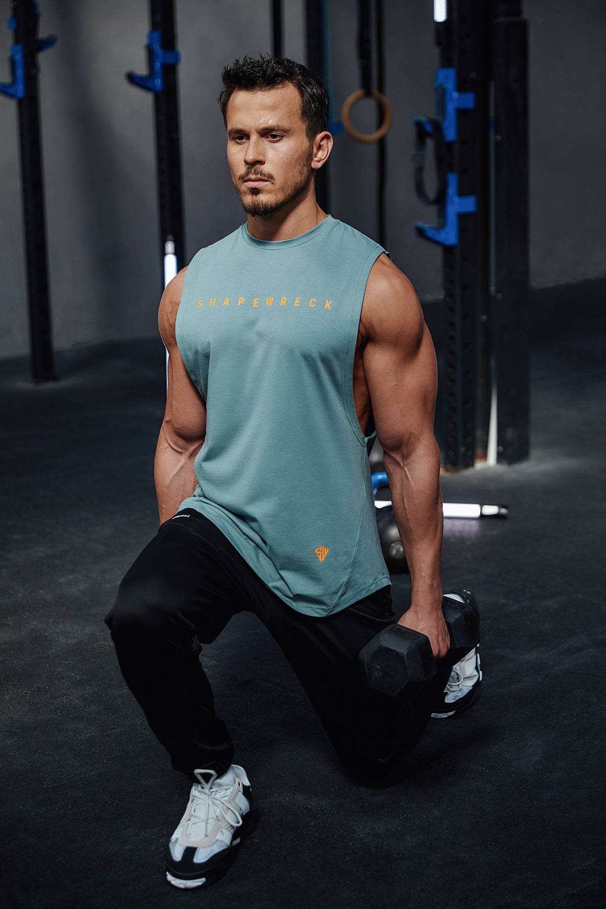 Man in a gym wearing a teal tank top and black pants, holding a dumbbell.